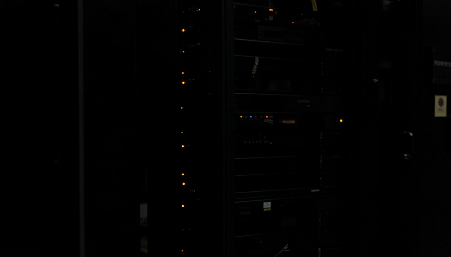 Dark photograph of a server rack in a dimly lit data center, with faint amber indicator lights glowing against deep shadow